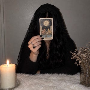May include: A tarot card titled The Moon featuring an illustration of a moon, two towers, a lobster, and two canines. The card is held up by a hand with black nail polish and silver rings, partially obscuring a face covered by a black lace veil.