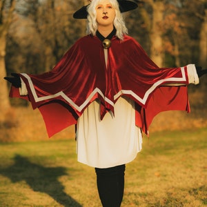 Pu&ograve; includere: Una donna con un vestito bianco e un mantello rosso con bordi bianchi si trova in un campo erboso. Indossa un cappello nero con orecchie da gatto e un campanello dorato intorno al collo. Il sole splende luminoso sullo sfondo.