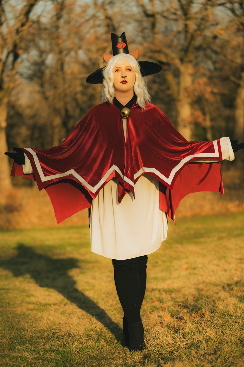 Pu&ograve; includere: Una donna con un vestito bianco e un mantello rosso con bordi bianchi si trova in un campo erboso. Indossa un cappello nero con orecchie da gatto e un campanello dorato intorno al collo. Il sole splende luminoso sullo sfondo.