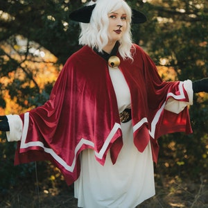 Pu&ograve; includere: Una donna con un vestito bianco e un mantello rosso con bordi bianchi, che indossa un cappello nero a forma di gatto con un solo occhio rosso. &Egrave; in piedi in un'area erbosa con alberi sullo sfondo.