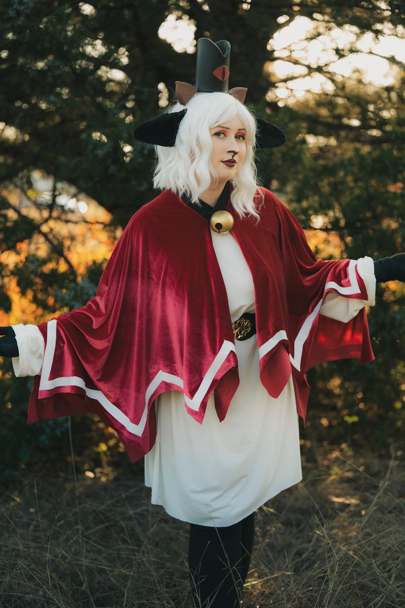 Pu&ograve; includere: Una donna con un vestito bianco e un mantello rosso con bordi bianchi, che indossa un cappello nero a forma di gatto con un solo occhio rosso. &Egrave; in piedi in un'area erbosa con alberi sullo sfondo.