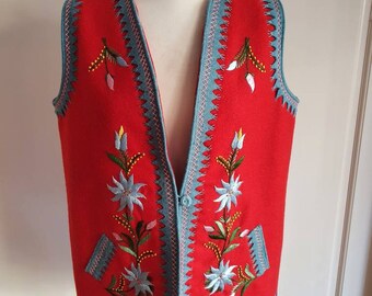 Pode incluir: Um colete de lã vermelho com um design floral bordado em azul e rosa. O colete tem um decote em V e um fecho de botão.