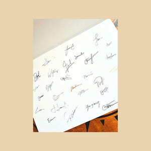 May include: A white sheet filled with numerous signatures in black ink. The signatures vary in style and size, with some appearing more elaborate than others. A few signatures are in red ink. The sheet is set against a neutral background.
