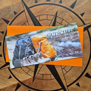 Könnte beinhalten: Ein orangefarbener Gutschein mit dem deutschen Wort "Gutschein" und den Worten "Wandertrip" aufgedruckt. Der Gutschein zeigt Bilder von Wanderschuhen und Rucksäcken. Der Gutschein befindet sich in einem orangefarbenen Umschlag, der auf einer hölzernen Windrose liegt.