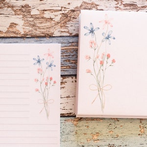 Puede incluir: Un juego de papeler&iacute;a con un bloc de notas rosa claro y un diario a juego. Ambas piezas est&aacute;n adornadas con un dise&ntilde;o floral en acuarela en tonos azules, rosas y melocot&oacute;n, atado con un delicado lazo. El bloc de notas tiene p&aacute;ginas rayadas.