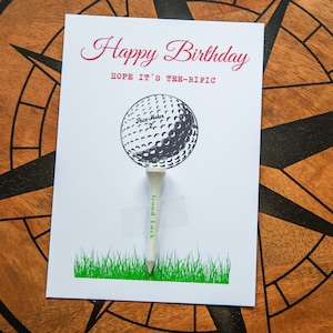 Könnte beinhalten: Eine weiße Geburtstagskarte mit Golf-Motiv. Die Karte zeigt den Text "Happy Birthday" in roter Schrift, eine Golfball-Illustration und ein Golf-Tee mit dem Aufdruck "Good Luck". Am unteren Rand ist grünes Gras aufgedruckt.