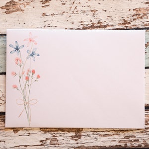 Puede incluir: Un sobre rosa claro con un dise&ntilde;o floral en acuarela. El dise&ntilde;o presenta flores azules y rosas con tallos verdes atados con un lazo de cuerda. El sobre est&aacute; en blanco, con un fondo de madera r&uacute;stica.