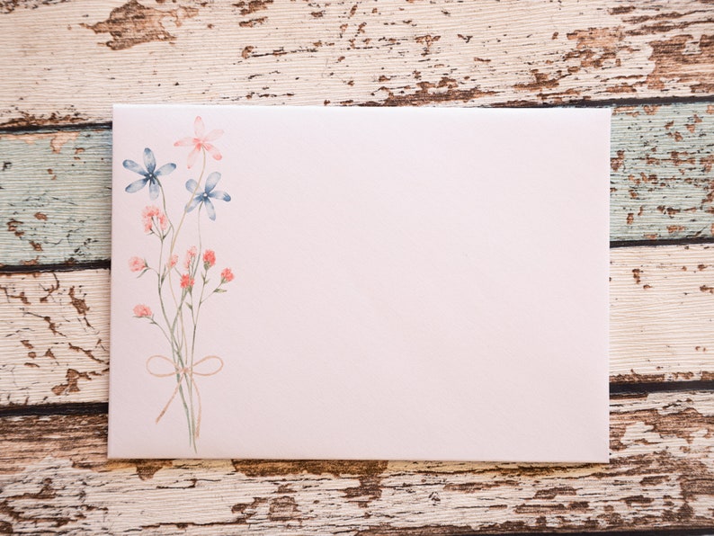 Puede incluir: Un sobre rosa claro con un dise&ntilde;o floral en acuarela. El dise&ntilde;o presenta flores azules y rosas con tallos verdes atados con un lazo de cuerda. El sobre est&aacute; en blanco, con un fondo de madera r&uacute;stica.