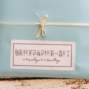 Puede incluir: Un juego de papeler&iacute;a azul claro atado con una cinta crema. La etiqueta rectangular indica "BRIEFPAPIER-SET" y contiene seis hojas de papel y seis sobres. La cinta est&aacute; atada con un lazo.