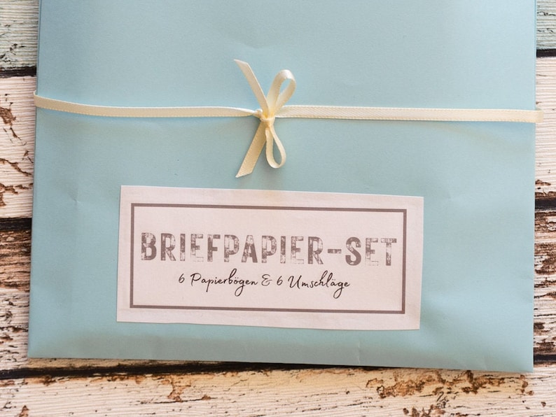 Puede incluir: Un juego de papeler&iacute;a azul claro atado con una cinta crema. La etiqueta rectangular indica "BRIEFPAPIER-SET" y contiene seis hojas de papel y seis sobres. La cinta est&aacute; atada con un lazo.
