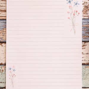 Puede incluir: Papel de papeler&iacute;a rayado con ilustraciones florales. El papel rosa claro tiene l&iacute;neas horizontales para escribir. Delicadas flores de estilo acuarela en rosa y azul est&aacute;n dispuestas en las esquinas superior derecha e inferior izquierda. El papel est&aacute; sobre una superficie de madera.