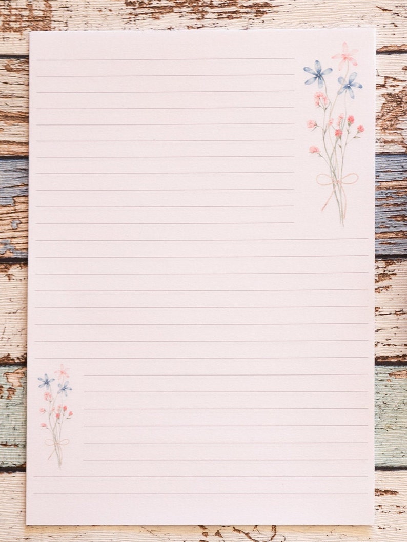 Puede incluir: Papel de papeler&iacute;a rayado con ilustraciones florales. El papel rosa claro tiene l&iacute;neas horizontales para escribir. Delicadas flores de estilo acuarela en rosa y azul est&aacute;n dispuestas en las esquinas superior derecha e inferior izquierda. El papel est&aacute; sobre una superficie de madera.