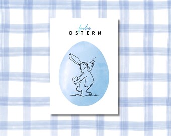May include: A greeting card with a blue watercolor egg design and a black and white bunny illustration. The words "Frohe Ostern" are written in German. The background is a blue and white plaid pattern.