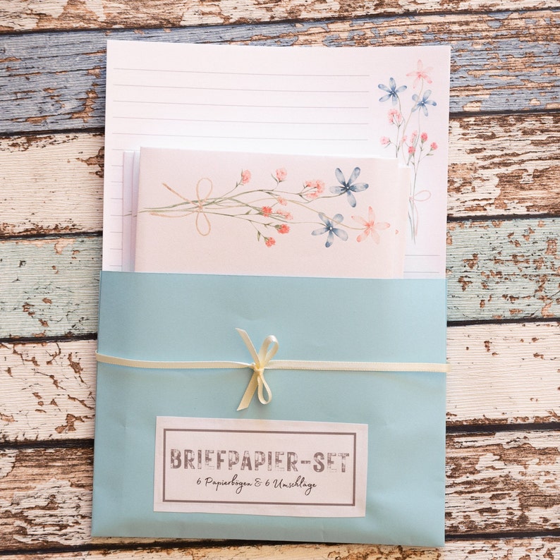 Puede incluir: Un juego de papeler&iacute;a con papel rayado, sobres y un soporte azul claro atado con una cinta crema. La papeler&iacute;a est&aacute; decorada con dise&ntilde;os florales de acuarela en rosa y azul. La etiqueta dice "BRIEFPAPIER-SET 6 Papierbogen & 6 Umschl&auml;ge."