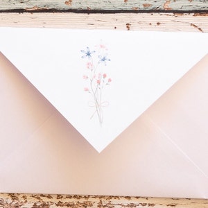 Puede incluir: Un sobre rosa claro con una solapa blanca con un dise&ntilde;o floral en acuarela. El dise&ntilde;o incluye flores azules y rosas atadas con un lazo delicado. El sobre est&aacute; sobre una superficie de madera r&uacute;stica.