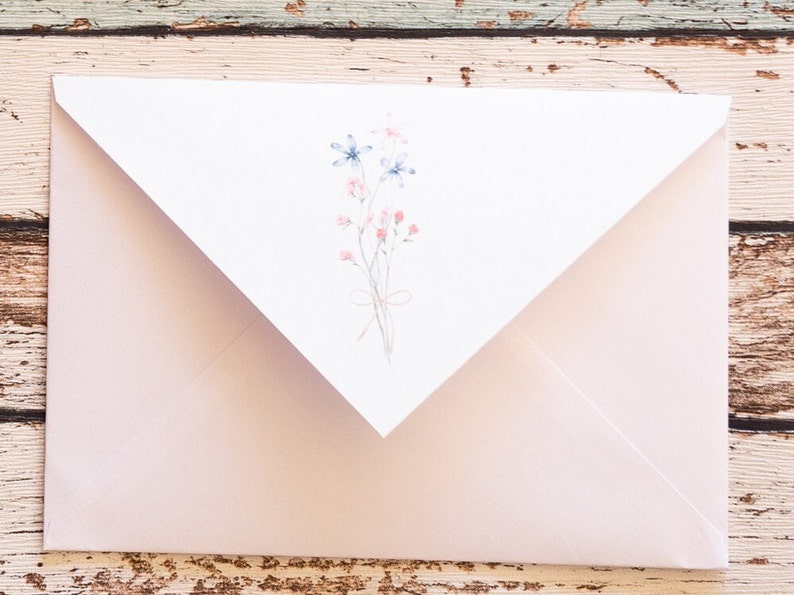 Puede incluir: Un sobre rosa claro con una solapa blanca con un dise&ntilde;o floral en acuarela. El dise&ntilde;o incluye flores azules y rosas atadas con un lazo delicado. El sobre est&aacute; sobre una superficie de madera r&uacute;stica.