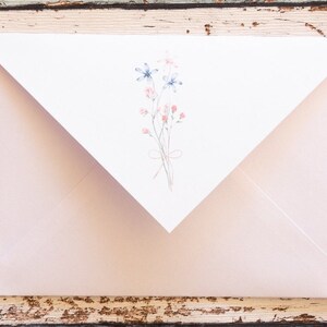 Puede incluir: Un sobre rosa claro con una solapa blanca con un dise&ntilde;o floral en acuarela. La ilustraci&oacute;n incluye flores azules y rosas atadas con un lazo delicado. El sobre est&aacute; sobre una superficie de madera r&uacute;stica y desgastada.