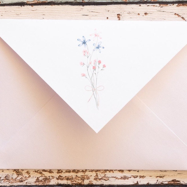 Puede incluir: Un sobre rosa claro con una solapa blanca con un dise&ntilde;o floral en acuarela. La ilustraci&oacute;n incluye flores azules y rosas atadas con un lazo delicado. El sobre est&aacute; sobre una superficie de madera r&uacute;stica y desgastada.