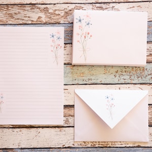 Puede incluir: Un juego de papeler&iacute;a con un bloc de notas rayado, sobres y tarjetas en blanco. Cada pieza es de color rosa suave con un delicado dise&ntilde;o floral en azul y coral. El conjunto est&aacute; dispuesto sobre una superficie de madera envejecida multicolor.