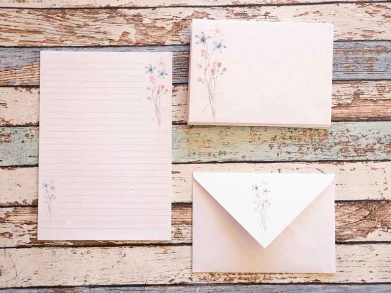 Puede incluir: Un juego de papeler&iacute;a con un bloc de notas rayado, sobres y tarjetas en blanco. Cada pieza es de color rosa suave con un delicado dise&ntilde;o floral en azul y coral. El conjunto est&aacute; dispuesto sobre una superficie de madera envejecida multicolor.