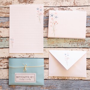 Puede incluir: Un juego de papeler&iacute;a con una hoja de escritura rayada, sobres y tarjetas. Los art&iacute;culos son de color rosa claro y blanco, adornados con ilustraciones florales en acuarela en azul y rosa. Tambi&eacute;n se incluye un sobre azul claro con la etiqueta "BRIEFPAPIER-SET".
