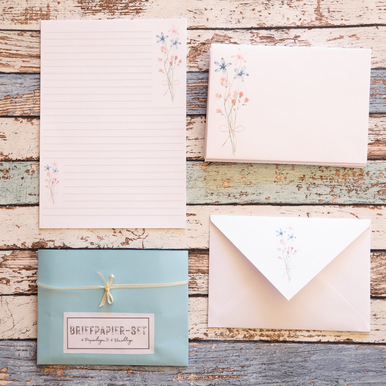 Puede incluir: Un juego de papeler&iacute;a con una hoja de escritura rayada, sobres y tarjetas. Los art&iacute;culos son de color rosa claro y blanco, adornados con ilustraciones florales en acuarela en azul y rosa. Tambi&eacute;n se incluye un sobre azul claro con la etiqueta "BRIEFPAPIER-SET".