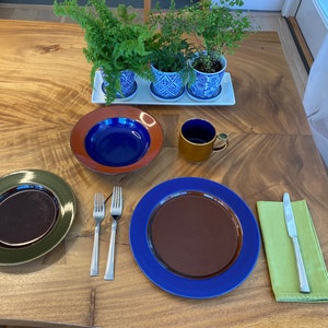 May include: A table setting with two place settings, each with a brown and green plate, a blue and brown plate, a silver fork, and a silver knife. A brown mug with a blue interior is on the table. Three potted plants are in the background.