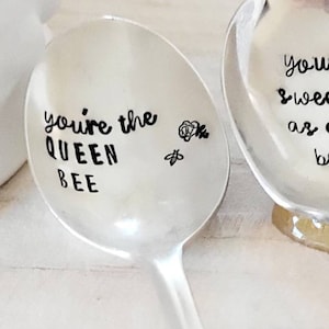 May include: Two silver spoons with engraved sayings. The first spoon says "you're the queen bee" with a small bee and flower design. The second spoon says "you're sweet as can bee" with a small bee and flower design.
