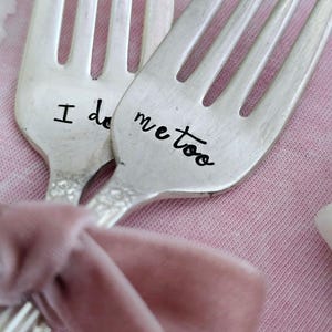 May include: Two silver forks with the words "I do" and "me too" stamped on the handles. A pink velvet ribbon is tied around the forks, resting on a pink fabric background. The forks are a unique wedding or engagement gift.