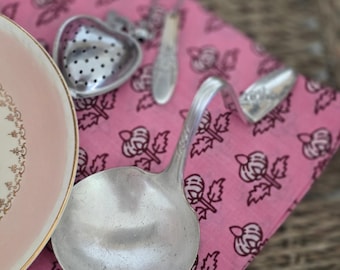 May include: A collection of vintage silverware and a heart-shaped tea infuser rest on a pink floral patterned cloth. A portion of a pink plate with a gold rim is visible. The items are arranged on a woven surface.