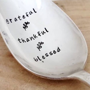 Op de afbeelding: Een zilveren lepel met de woorden "grateful", "thankful" en "blessed" erop gegraveerd. Elk woord wordt vergezeld door een klein blaadjesontwerp.