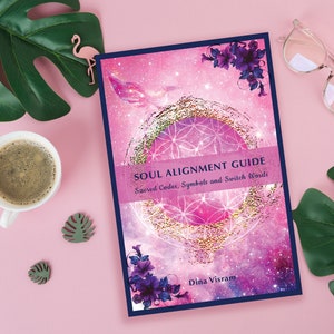 May include: A pink and purple book with a gold and white geometric design on the cover. The title is "Soul Alignment Guide: Sacred Codes, Symbols and Switch Words" by Dina Visram.