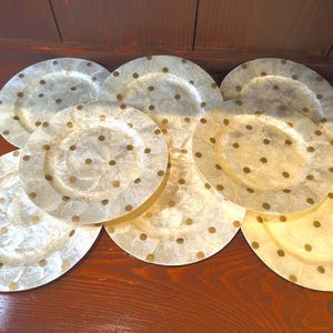 Puede incluir: Un conjunto de siete platos decorativos. Los platos son de color blanquecino con un brillo nacarado y están adornados con pequeños lunares dorados espaciados uniformemente. Los platos están colocados sobre una superficie de madera oscura.