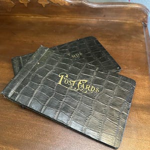 May include: A stack of black vintage postcard albums with gold lettering reading "Post Cards" on a wooden surface. The albums have a textured, crocodile-like pattern. The albums are open, revealing the interior pages.