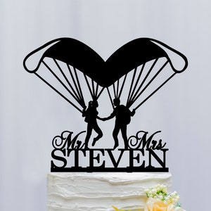 Könnte beinhalten: Schwarze Silhouette eines Paares, das sich beim Fallschirmspringen an den Händen hält, mit einem Fallschirm über ihnen. Der Text "Mr. & Mrs. STEVEN" befindet sich unter dem Paar. Der Tortendekor befindet sich auf einer weißen Hochzeitstorte.