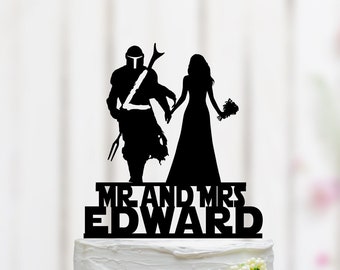 May include: Black silhouette cake topper of a couple, a bride and groom, with the groom dressed as a Mandalorian. The text "MR AND MRS EDWARD" is below the couple.
