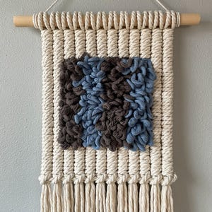 May include: A rectangular macrame wall hanging with a natural wood dowel and off-white cotton rope. The central square features textured blue and brown yarn. The bottom edge has long fringe.