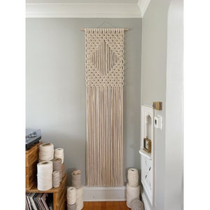 May include: A large, off-white macrame wall hanging with a geometric pattern. The hanging is made of natural fiber rope and has a wooden dowel at the top.