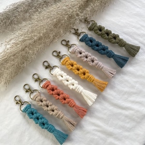 May include: A set of eight colorful macrame keychains with tassels. Each keychain features a double-knotted flower design in a different color, including teal, beige, orange, white, yellow, pink, and green.