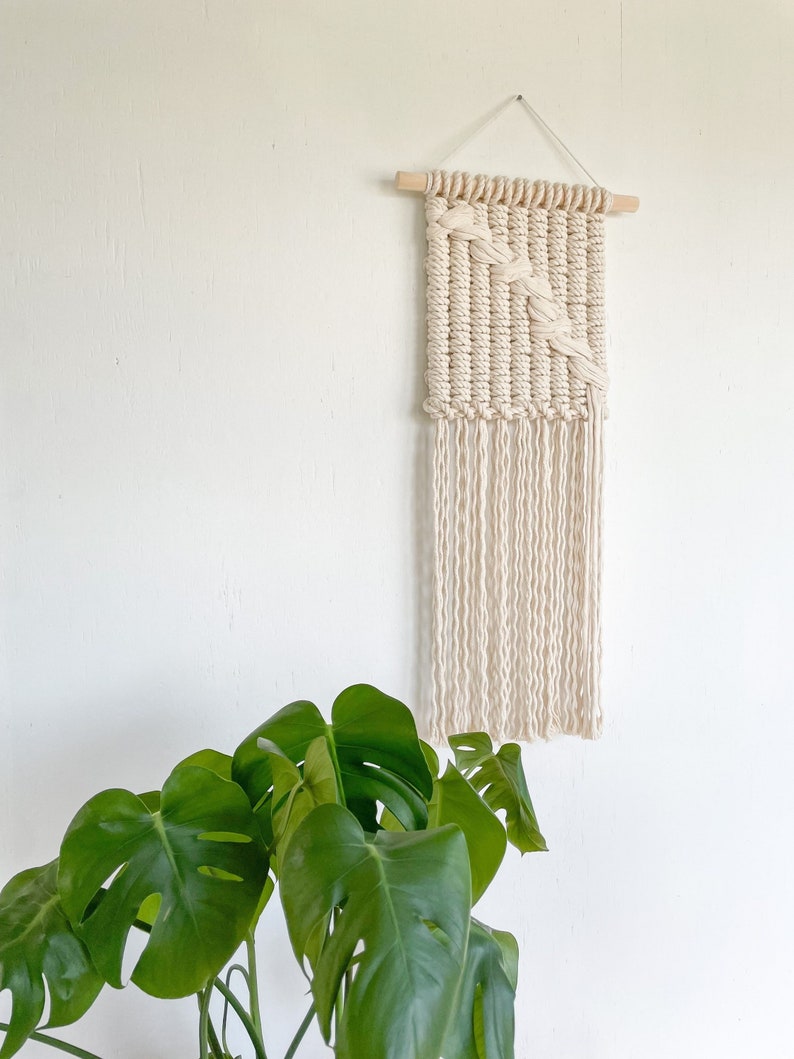 May include: A white macrame wall hanging with a geometric pattern and fringe. The hanging is made with natural cotton cord and is attached to a wooden dowel.