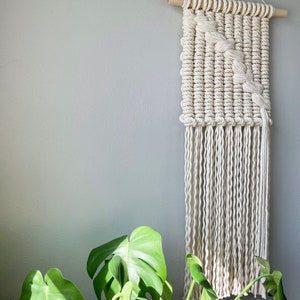May include: A white macrame wall hanging with a geometric pattern and fringe. The hanging is made with natural cotton rope and is mounted on a wooden dowel.