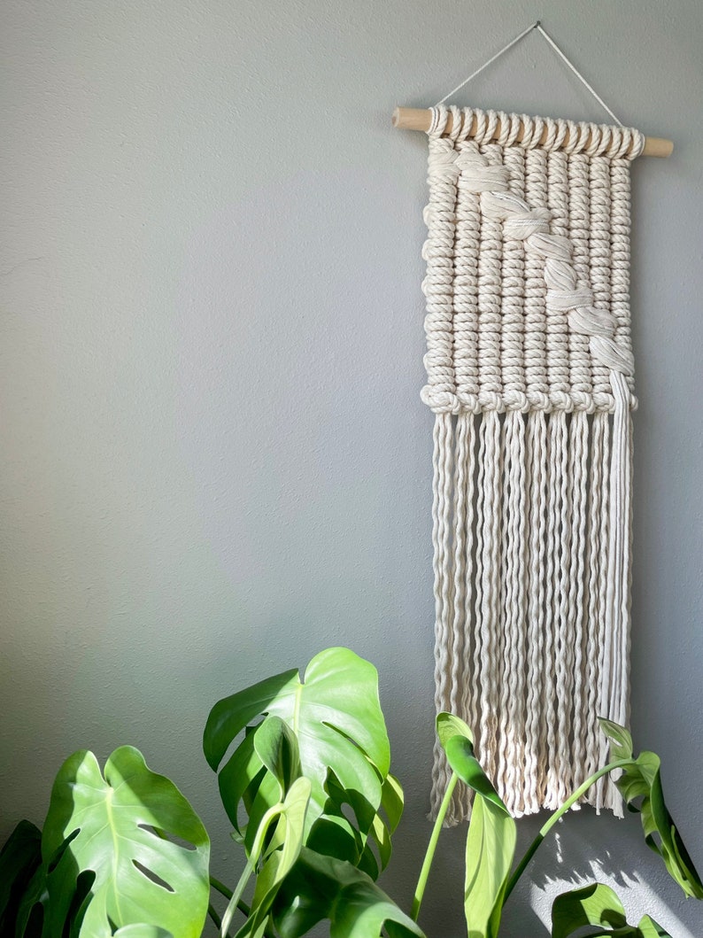 May include: A white macrame wall hanging with a geometric pattern and fringe. The hanging is made with natural cotton rope and is mounted on a wooden dowel.