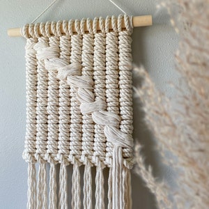 May include: A white macrame wall hanging with a diagonal pattern and fringe. The hanging is made with natural cotton rope and is attached to a wooden dowel.