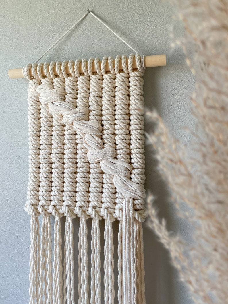 May include: A white macrame wall hanging with a diagonal pattern and fringe. The hanging is made with natural cotton rope and is attached to a wooden dowel.