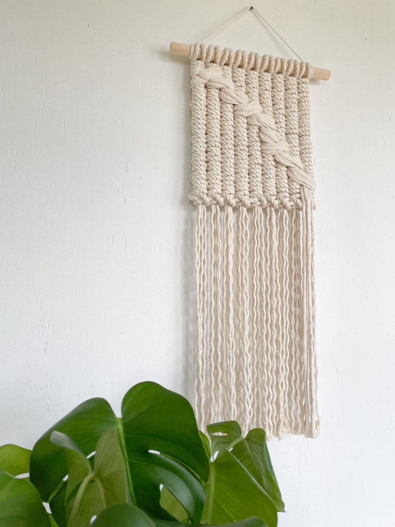 May include: A white macrame wall hanging with a geometric pattern and long fringe. The hanging is attached to a wooden dowel rod.