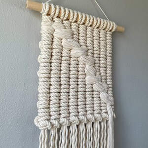 May include: A white macrame wall hanging with a geometric design and fringe. The hanging is made with natural cotton rope and is attached to a wooden dowel.