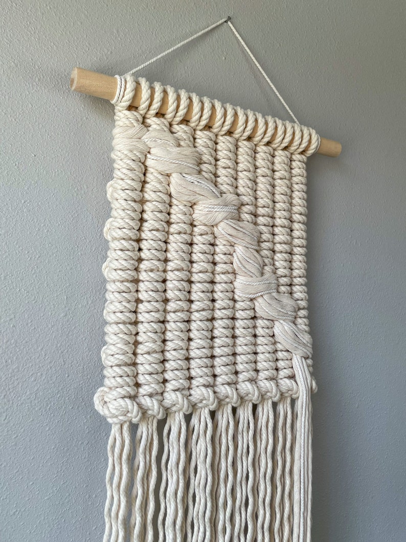 May include: A white macrame wall hanging with a geometric design and fringe. The hanging is made with natural cotton rope and is attached to a wooden dowel.