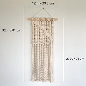 May include: A white macrame wall hanging with a geometric pattern and fringe. The hanging is 32 inches tall and 12 inches wide.
