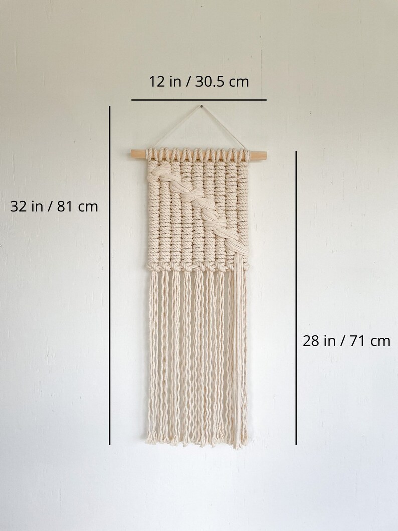 May include: A white macrame wall hanging with a geometric pattern and fringe. The hanging is 32 inches tall and 12 inches wide.
