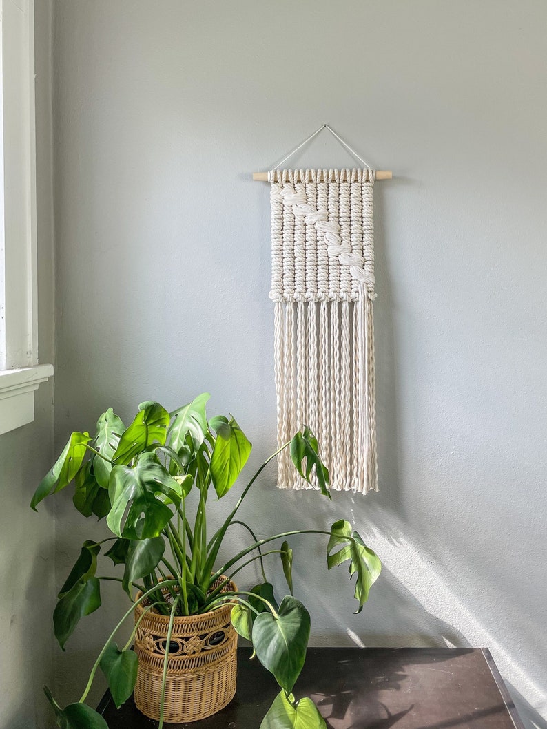 May include: A white macrame wall hanging with a geometric pattern and fringe. The hanging is mounted on a light wood dowel and is displayed against a light gray wall. A green potted plant is in the foreground.